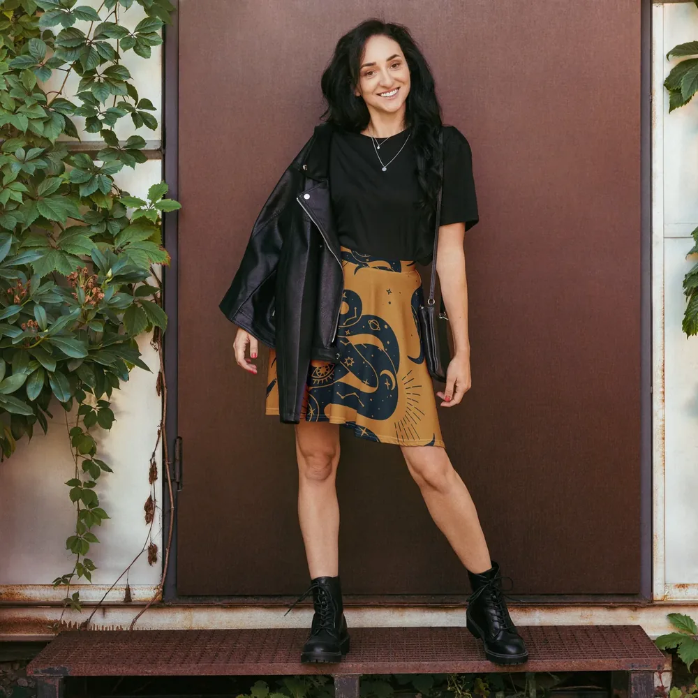 A woman wears the Arcana Skater Skirt, mustard with a navy serpent print, with a black tee and leather jacket in front of a dark door.