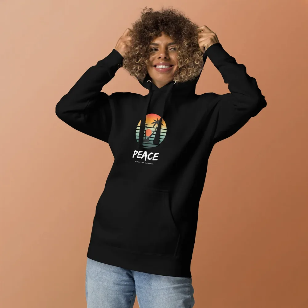 A woman wears the black Quiet Peace Hoodie, printed with a sunset palm and the word Peace, against a terracotta wall.
